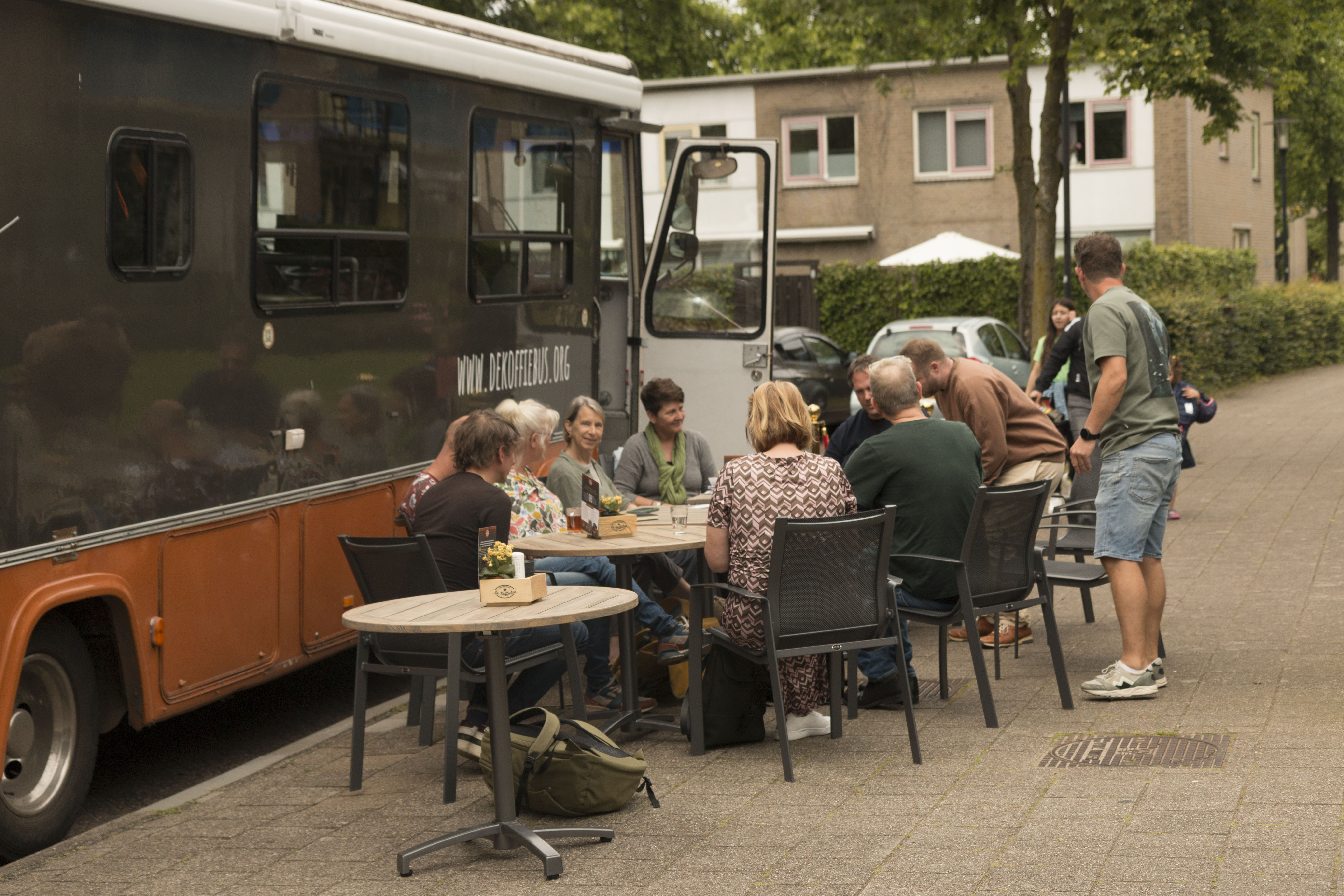 Foto van een mobiele ontmoetingsplek. Mensen zitten aan tafeltjes bij een busje.